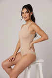 Woman wearing a beige bodysuit sitting on a white stool against a plain background